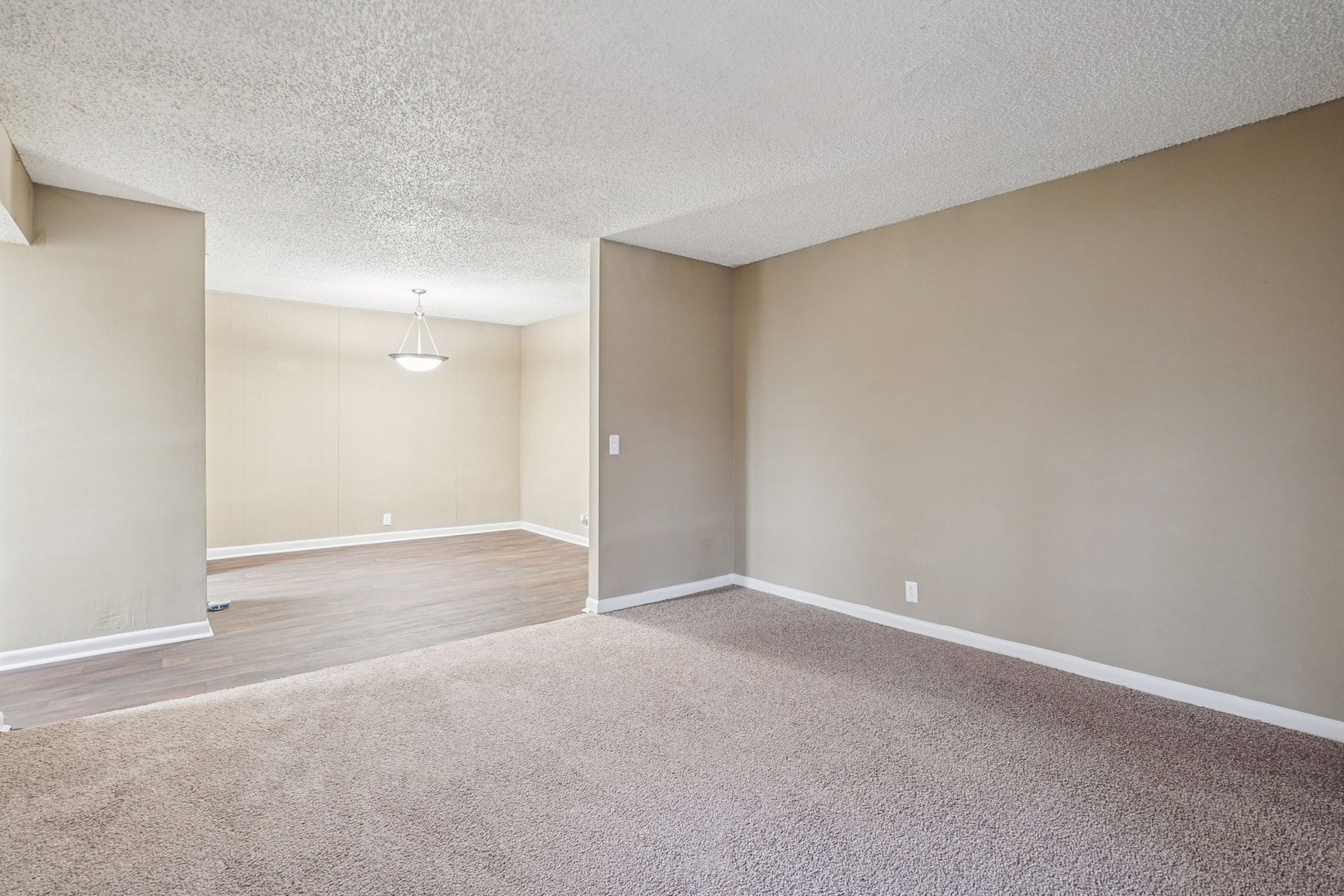 A vacant room with beige walls and gray carpeted flooring, featuring an open doorway leading to a dining area.