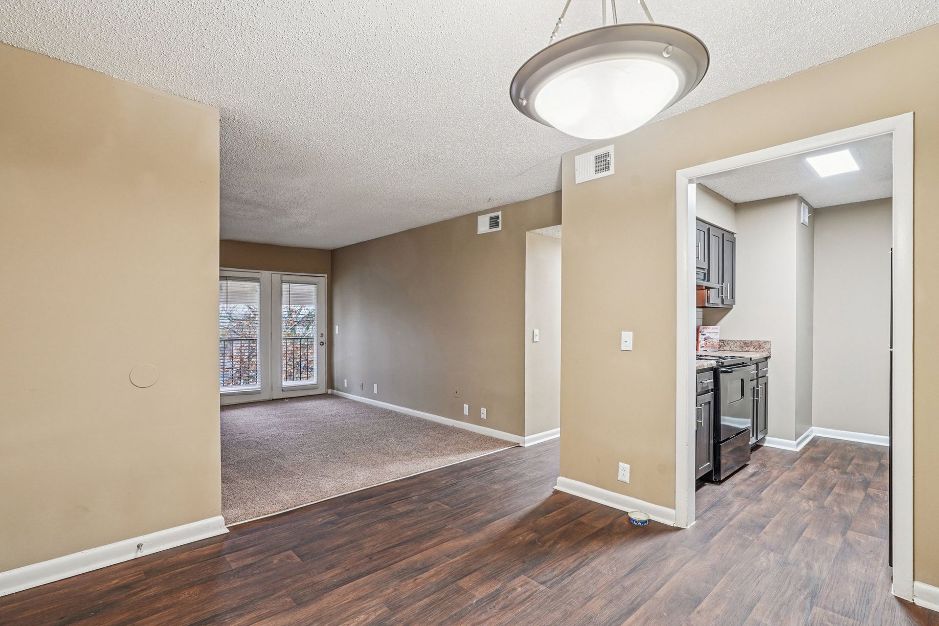 An interior view showing a dining area with wood-style flooring leading to a carpeted living room and a kitchen doorway.