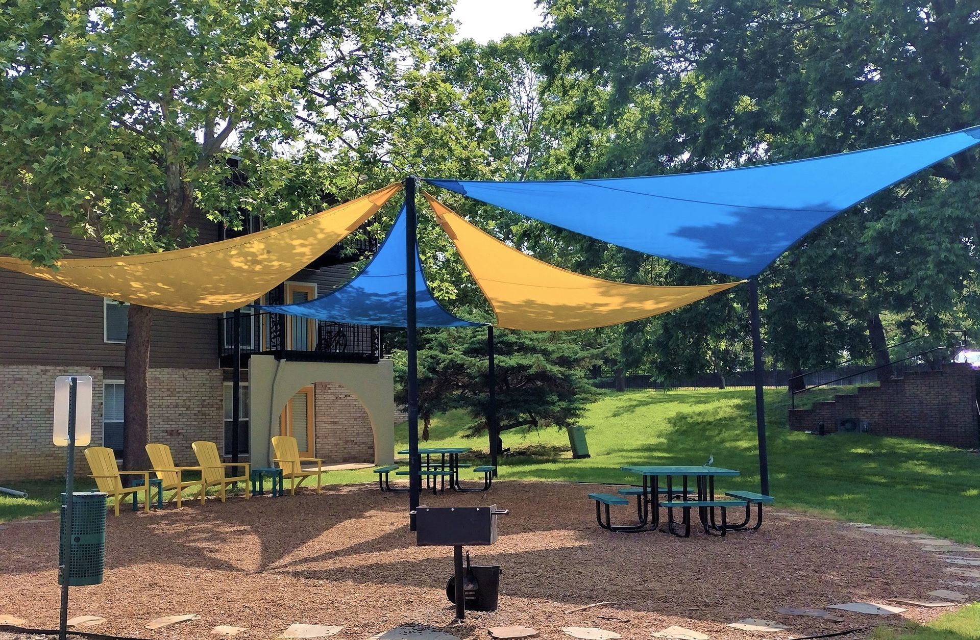 A park area with picnic tables and chairs under yellow and blue triangular sun shades on a mulch-covered ground.