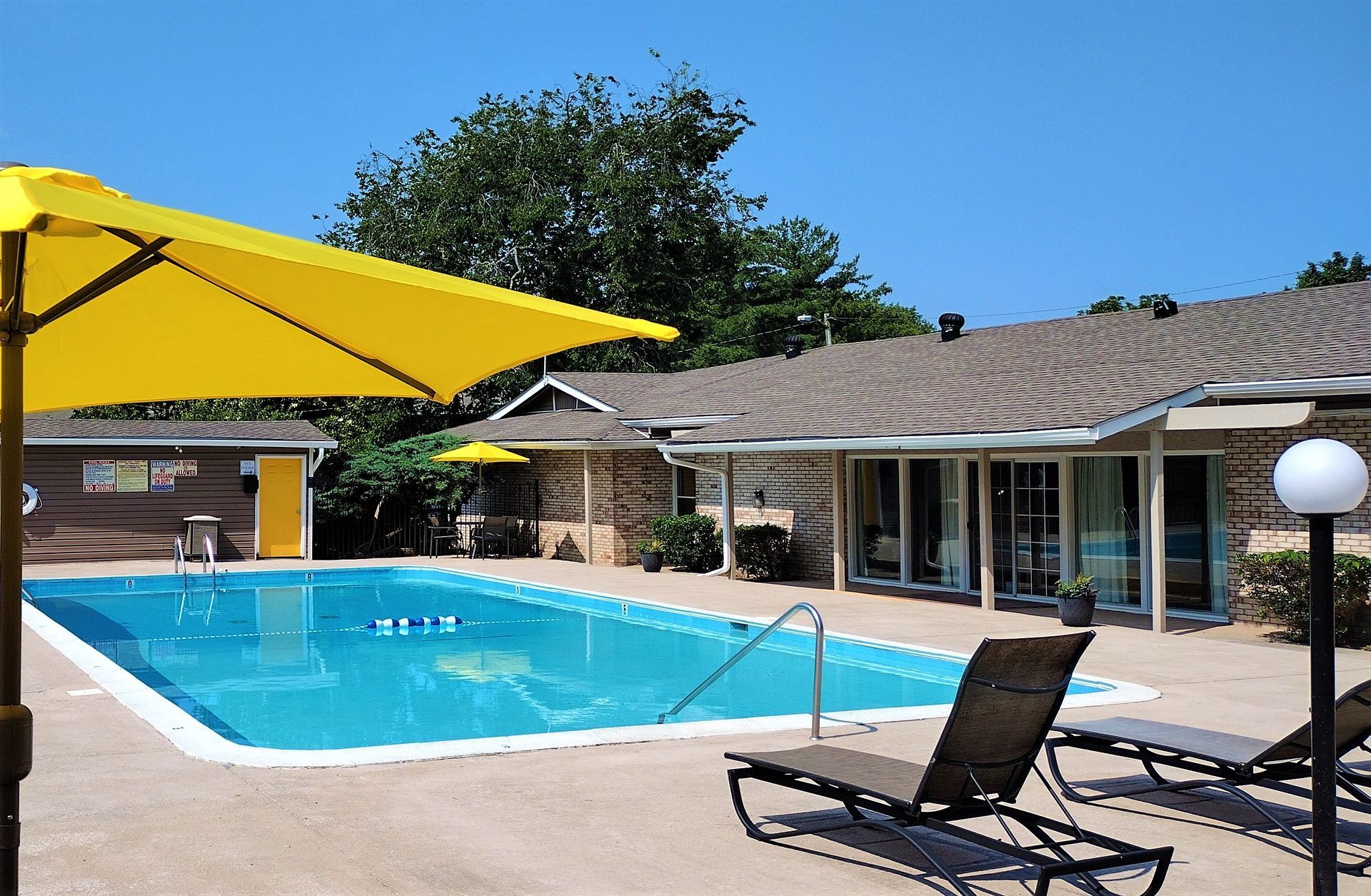 A rectangular swimming pool with bright blue water sits in front of a stone building, under a clear, sunny blue sky.