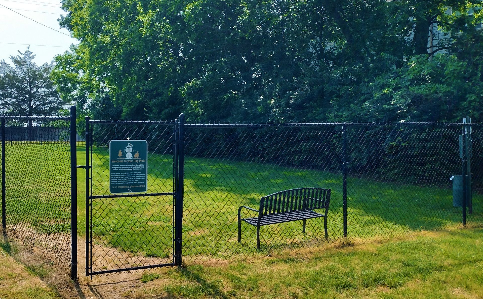 A grassy dog park area enclosed by a black chain-link fence, featuring a single metal bench and a gated entrance.