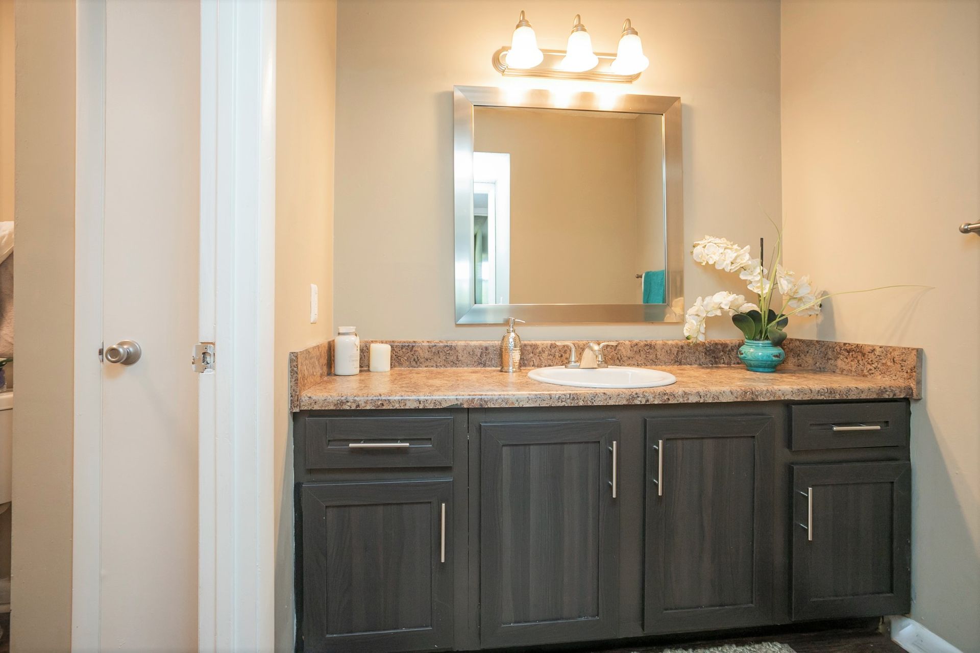 A bathroom vanity with dark gray cabinets, a speckled granite countertop, a mirror, and an orchid flower arrangement.