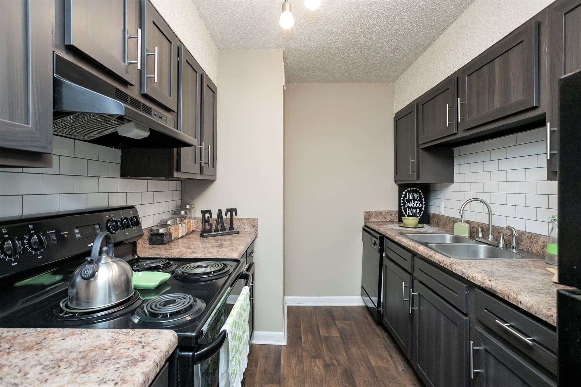 A modern galley-style kitchen featuring dark wood cabinets, speckled countertops, white subway tiles, and a tea kettle.