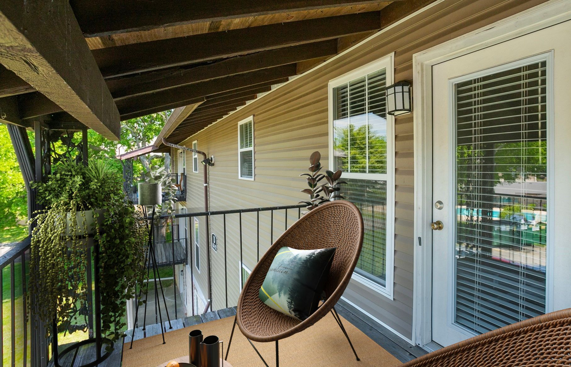 A tan balcony with a round wicker chair, a potted plant, and a glass door reflecting trees and a swimming pool.