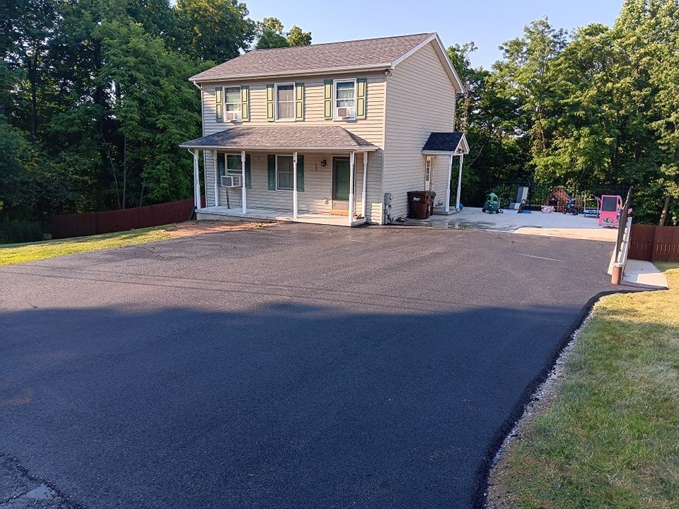 Two-story beige house with a newly paved black driveway. Green trees surround the property.
