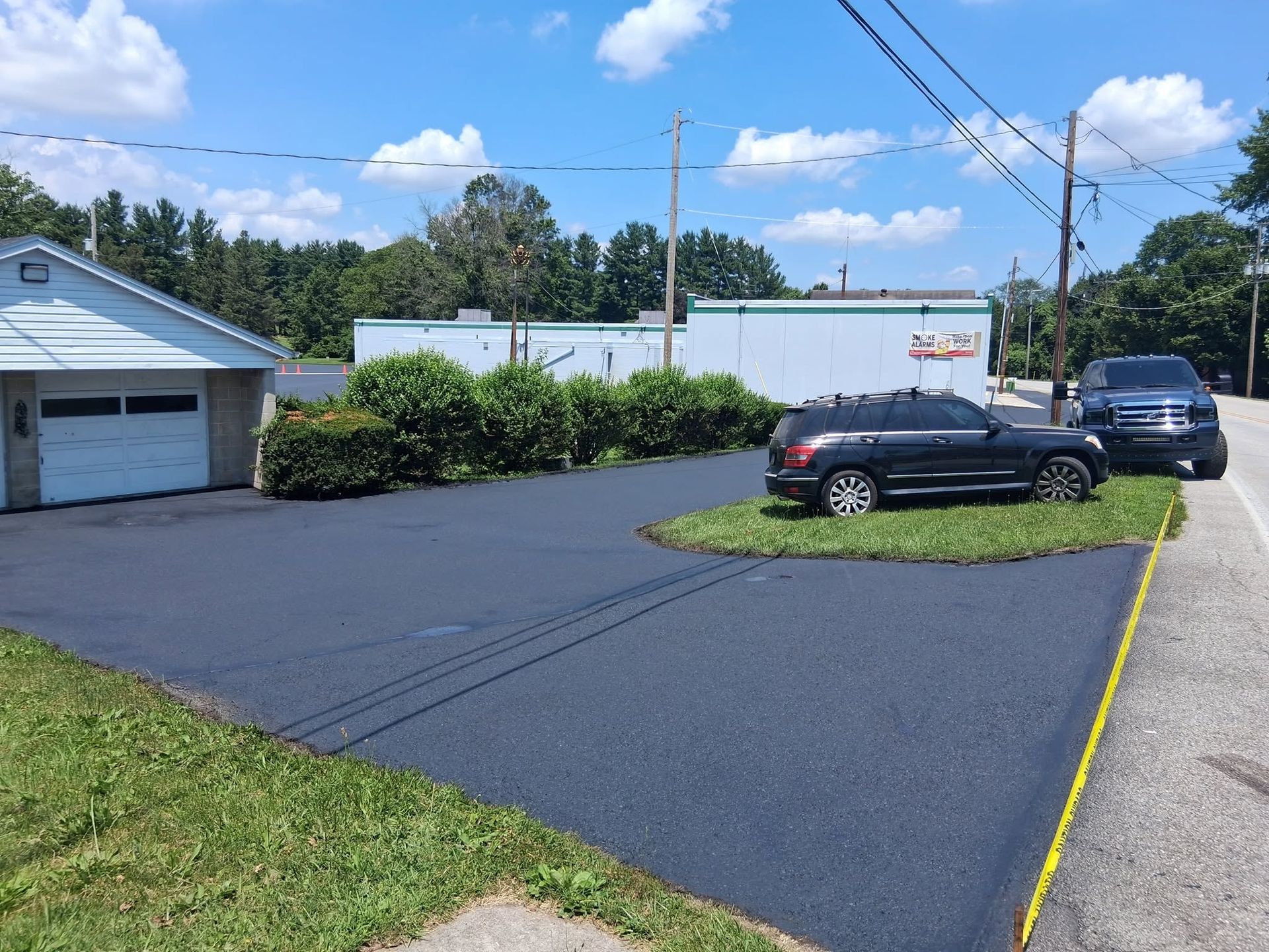 Paved driveway with a car parked on grass, next to a road, building with garage, and trees under a sunny sky.