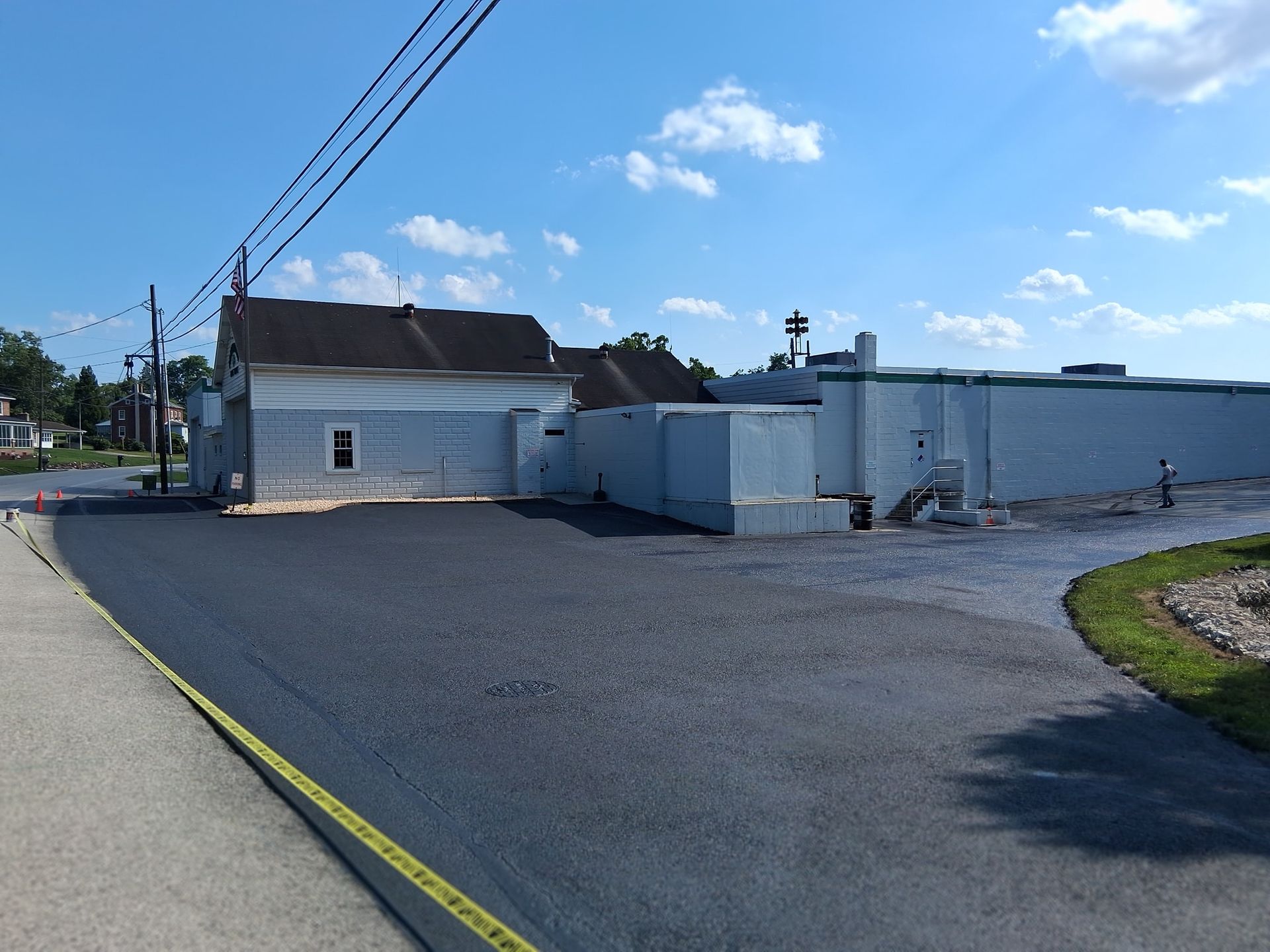 Paved parking lot in front of light blue building and older house. Power lines above. Sunny, blue sky.