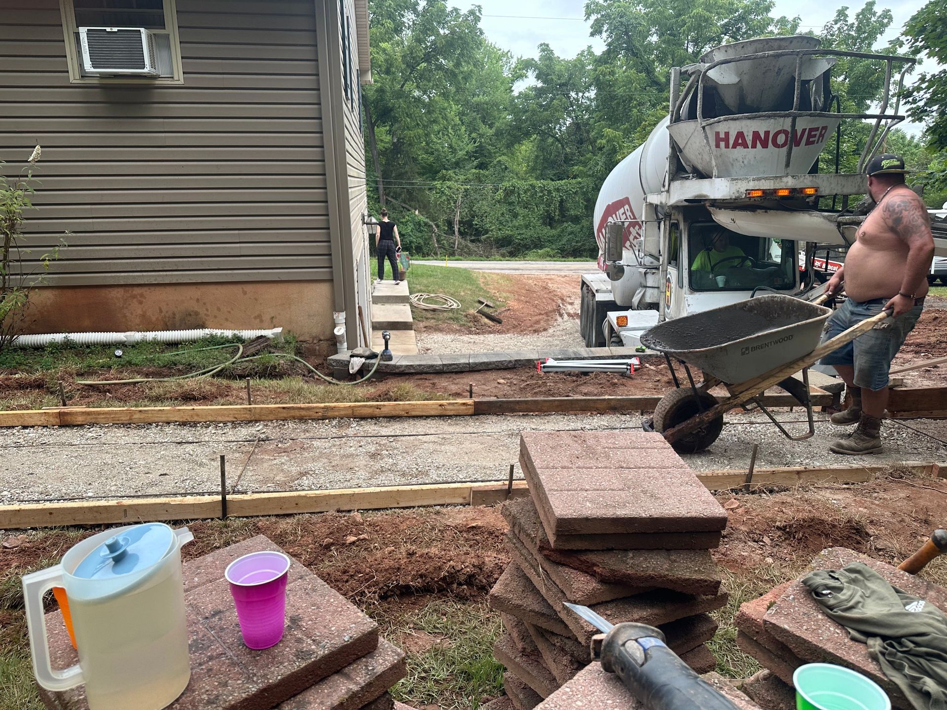 Concrete pour in progress: Workers, cement truck, wheelbarrow, and building. Brick pavers and drinks nearby.