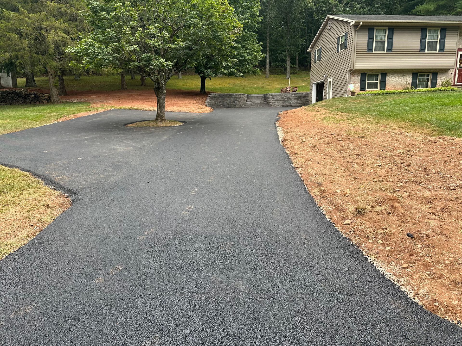 Asphalt driveway leading to a two-story house with green lawn. Dirt and a tree are present.