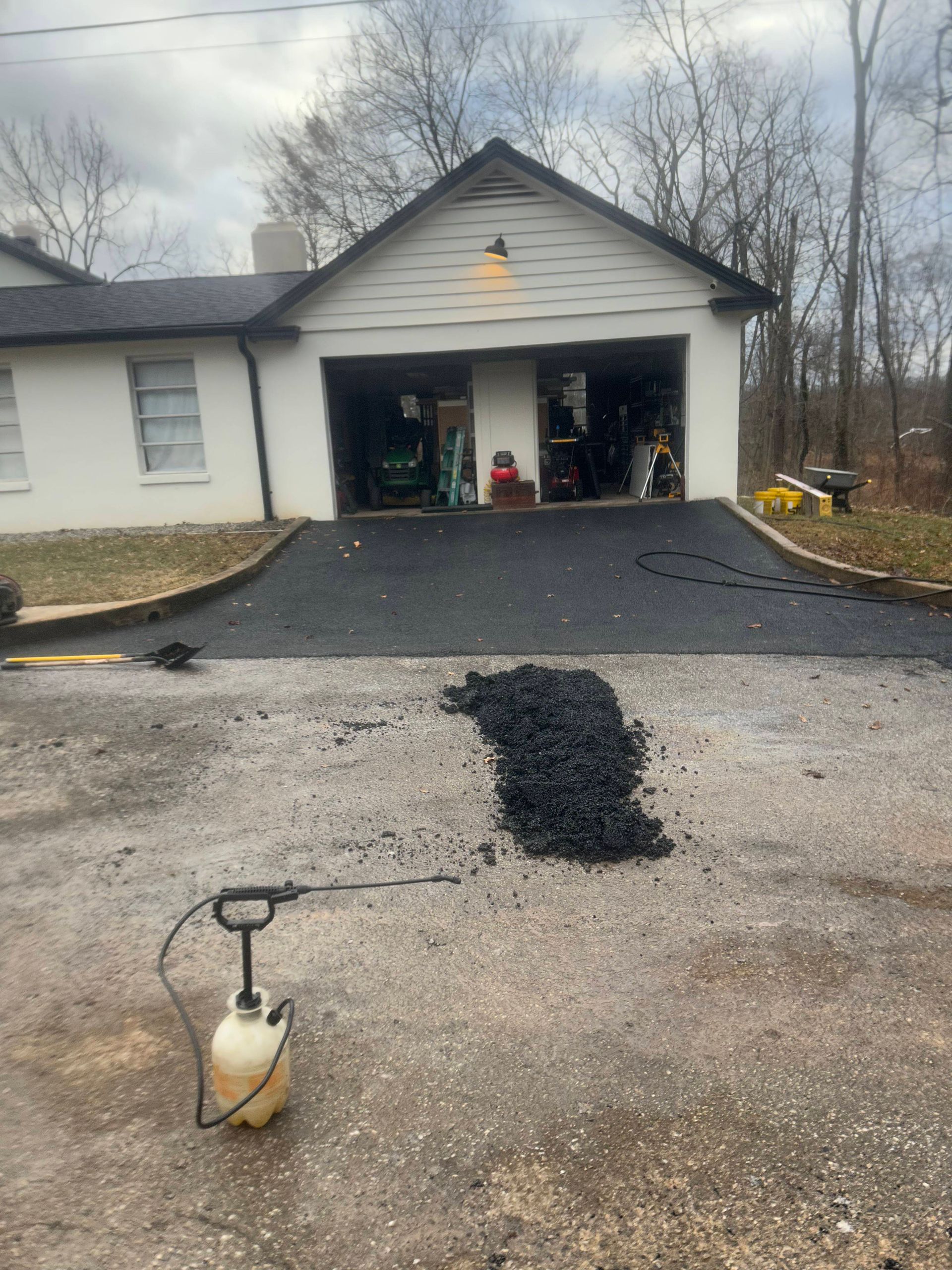 Asphalt driveway being repaired. Black asphalt pile with sprayer. White house and garage in background.