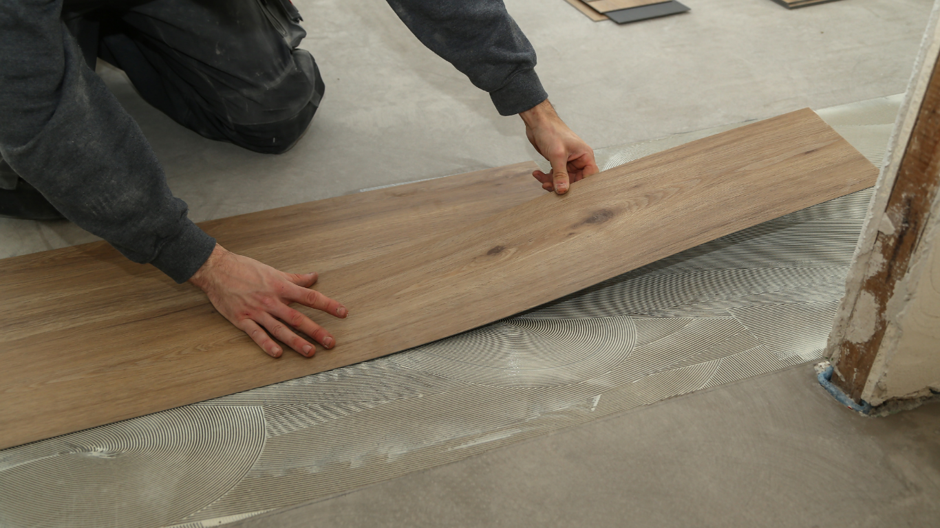 A person kneeling on a floor, carefully installing a wood-look plank onto a surface with spread adhesive.