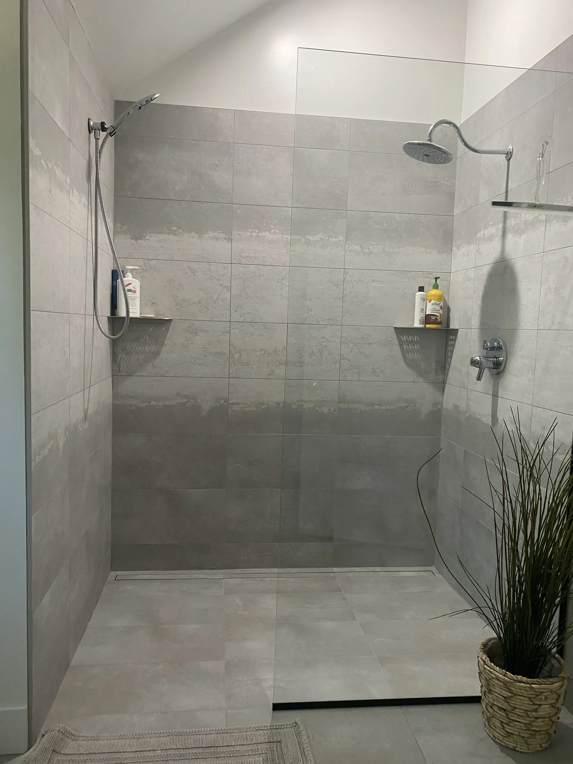 A modern walk-in shower with large grey tiles, two shower heads, corner shelves, and a glass partition.