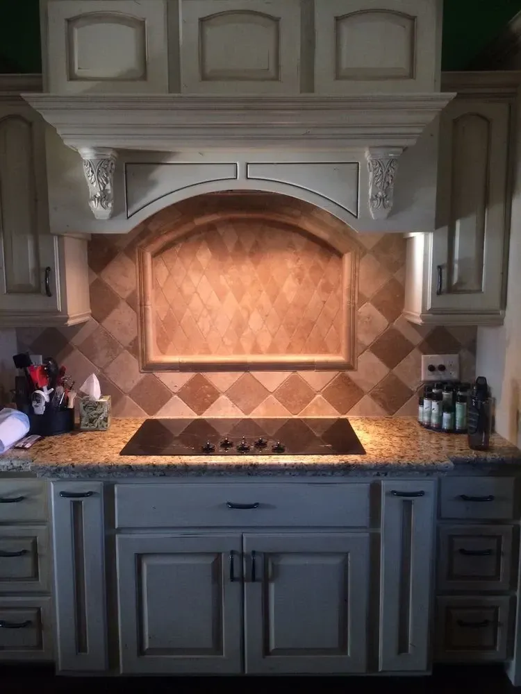 A kitchen stove area featuring light wood cabinets, a stone tile backsplash in a diamond pattern, and a granite countertop.