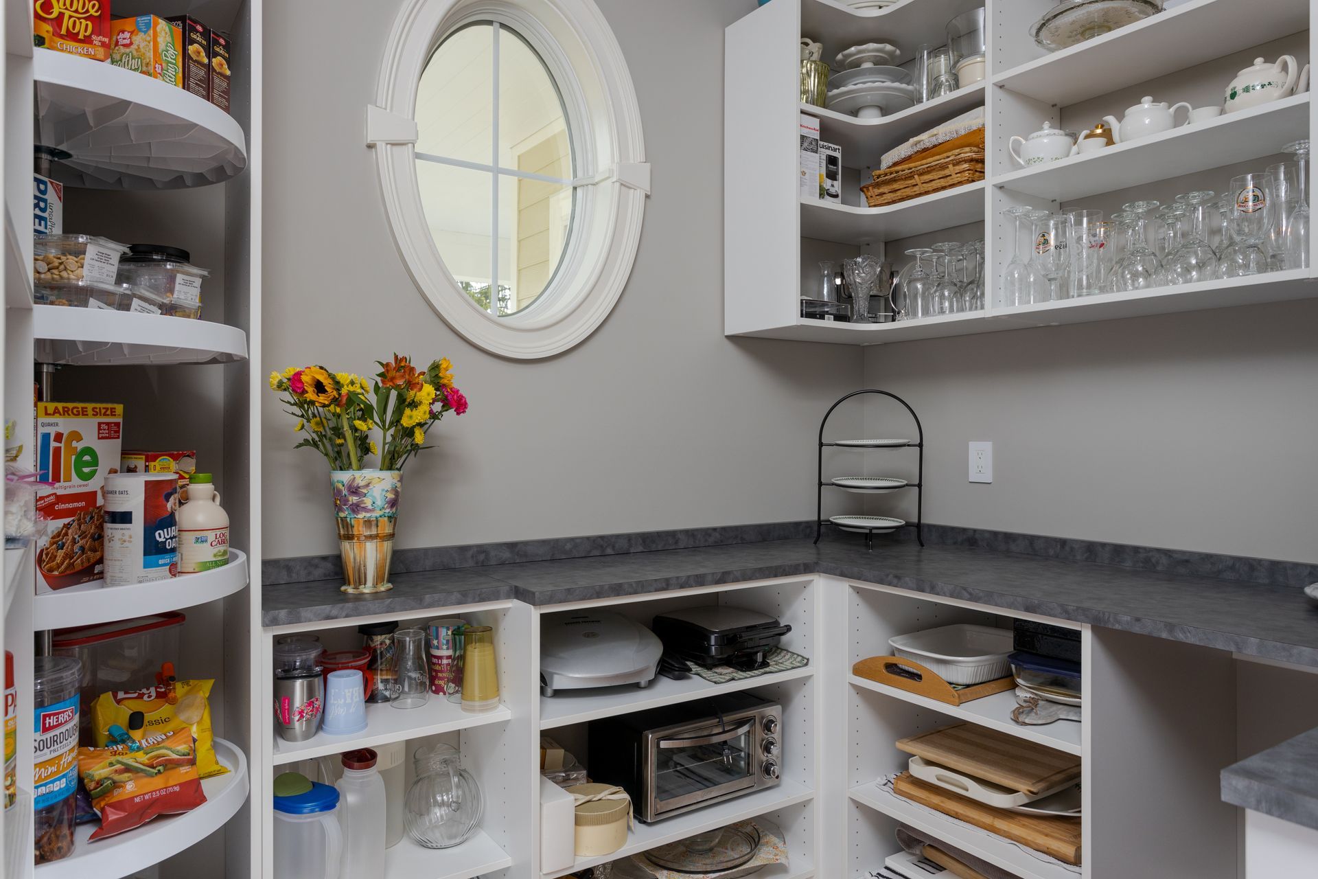 Pantry with white shelves, canned goods, glasses, and a vase of flowers beside a small round window.