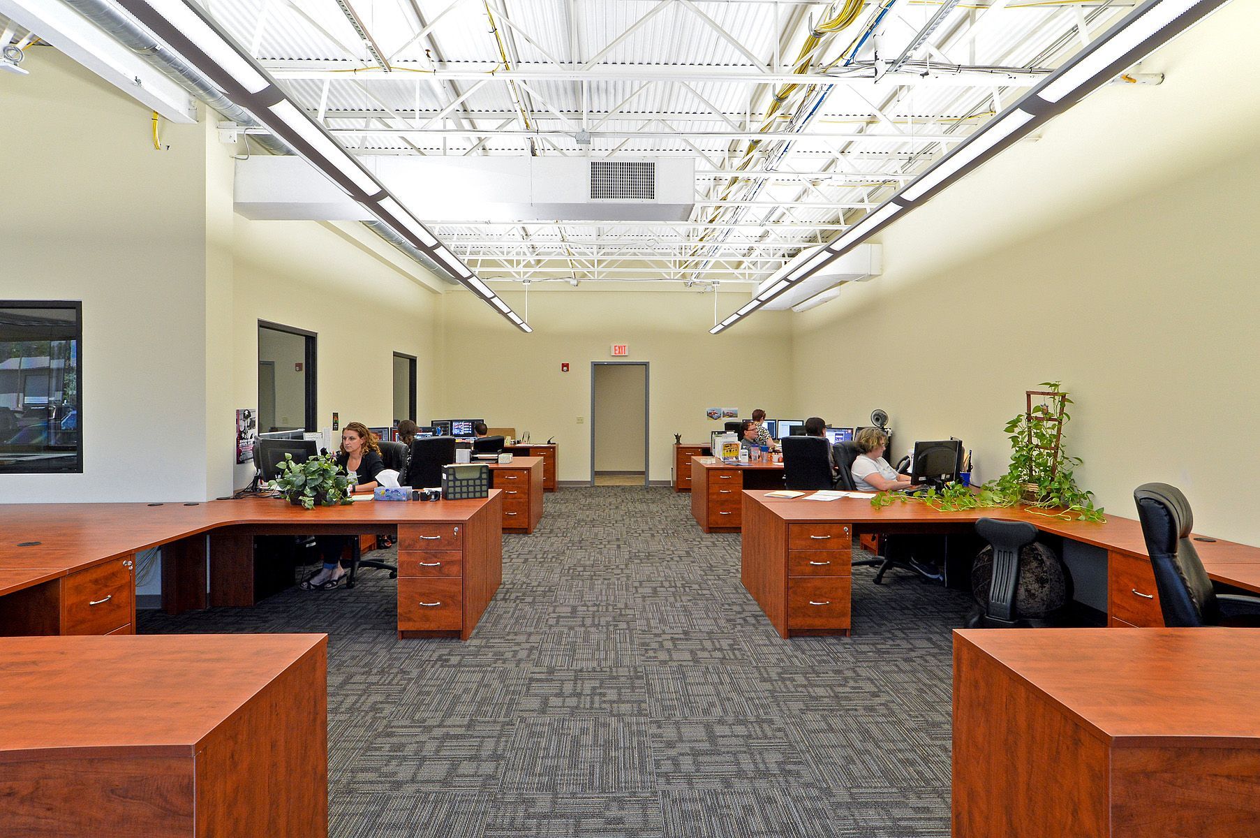 Open office with wooden desks, black chairs, and a few people working under bright ceiling lights.