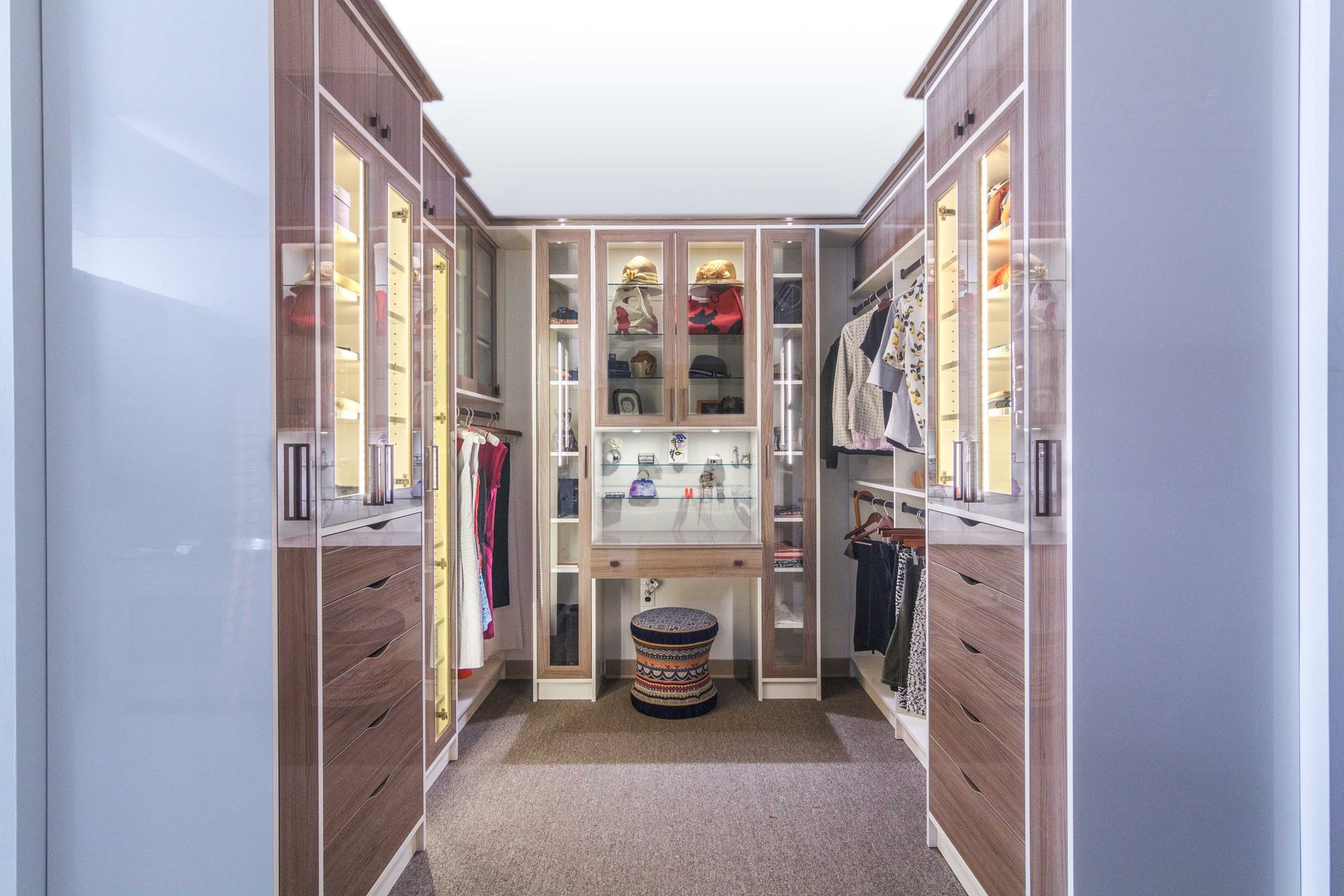 Bright walk-in closet with glass-front cabinets, hanging clothes, and a patterned stool in the center.