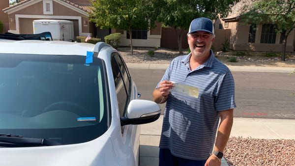 A man is standing in front of a white car holding a check.