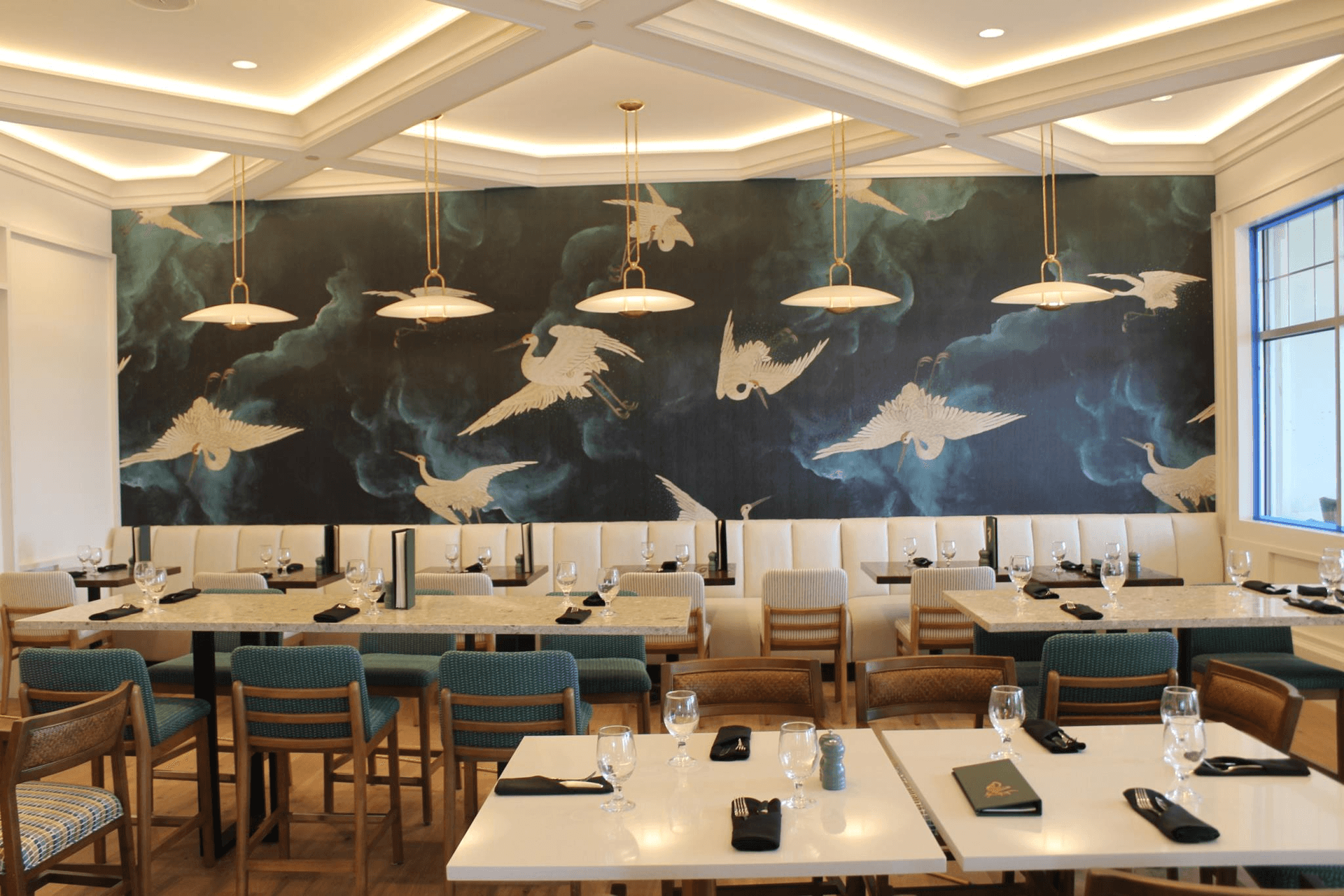 Restaurant interior with tables, blue chairs, white banquette, and bird mural.