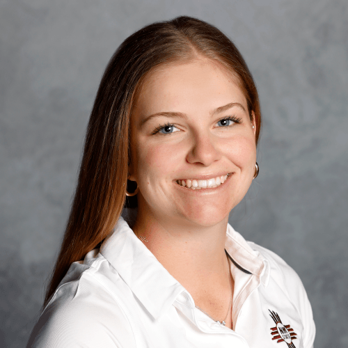 Tennis player in white shorts and visor swings racket on green court, shadow visible.