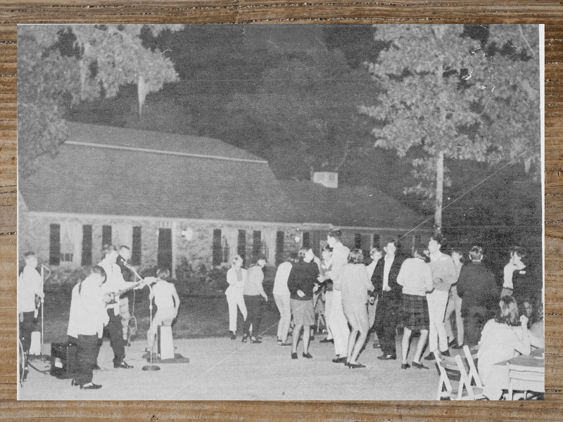A group of people gathers outdoors in front of a long building, possibly a social gathering or party.