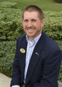 Tennis player in white shorts and visor swings racket on green court, shadow visible.