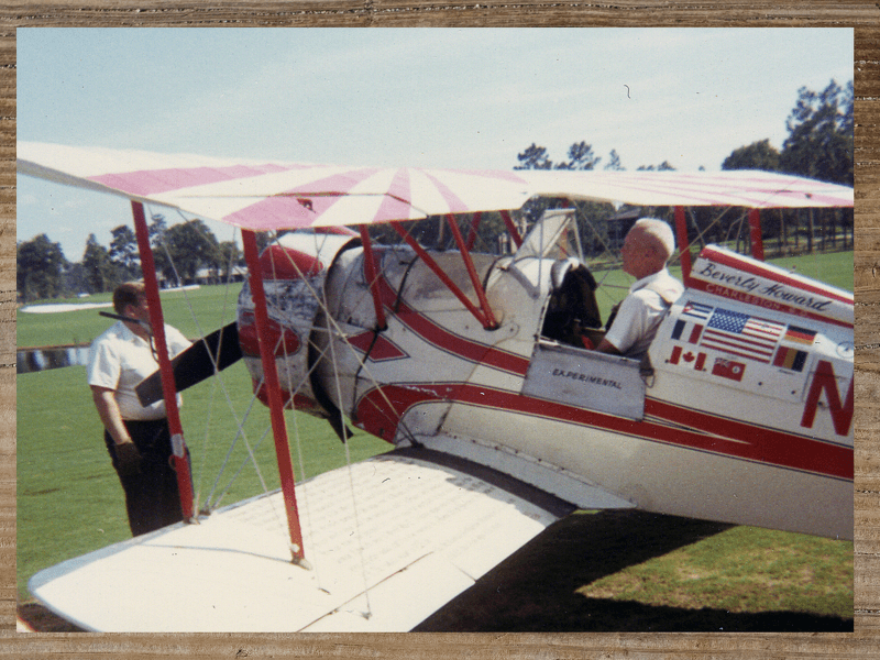 Biplane with red and white stripes; two men in shirtsleeves near the plane on grass. American flag on the plane.