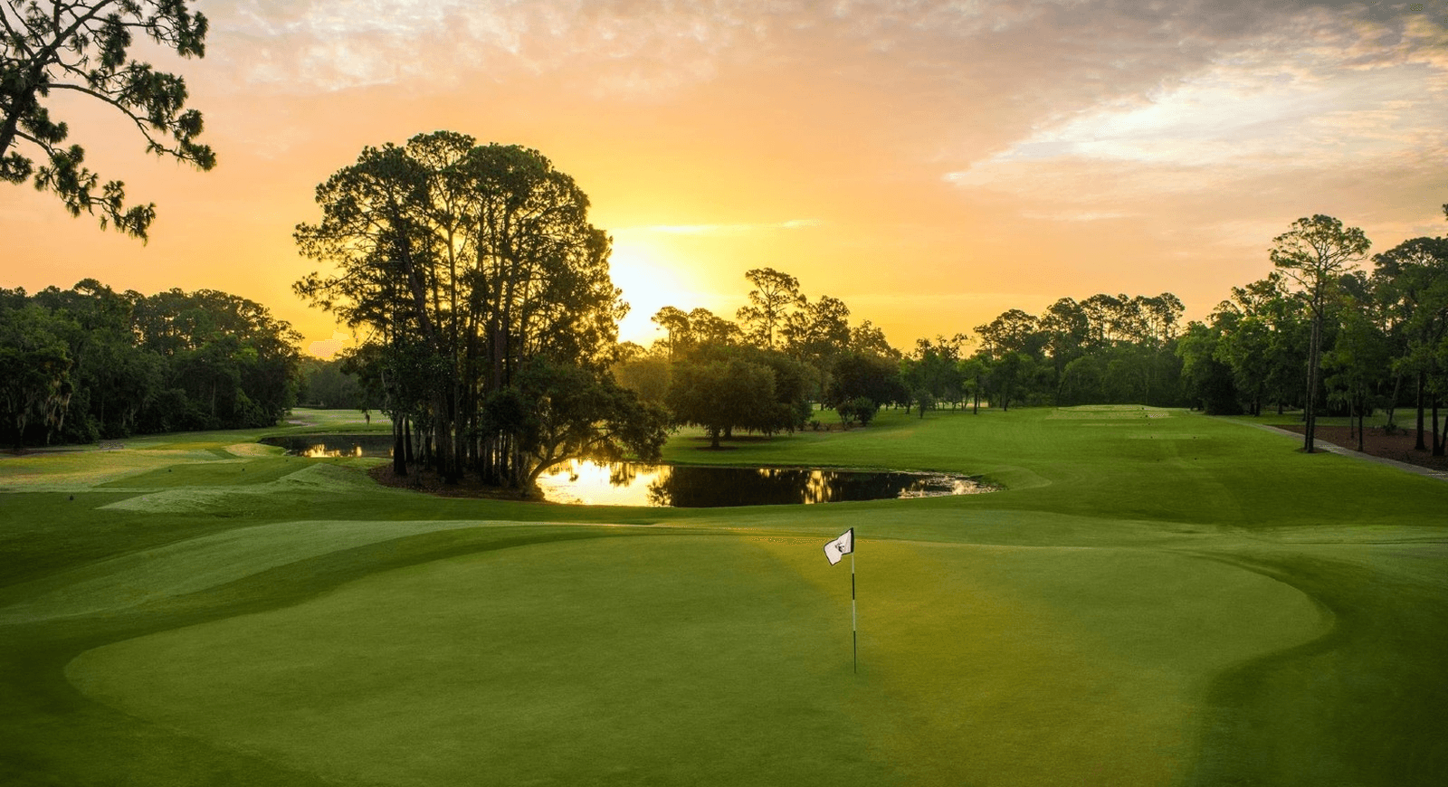 Golf course at sunset, green fairways and water feature, sun setting behind trees.