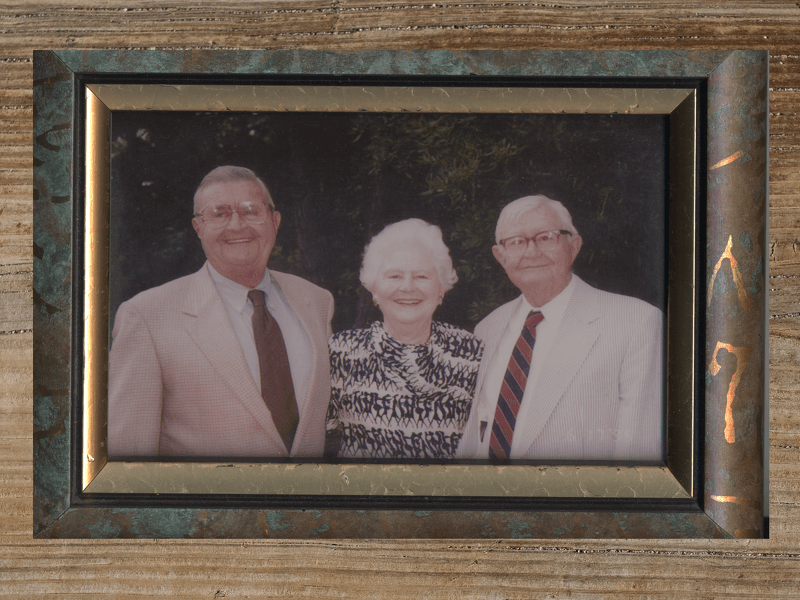 Three people in a framed portrait: two men and a woman, all wearing dress attire, smiling.