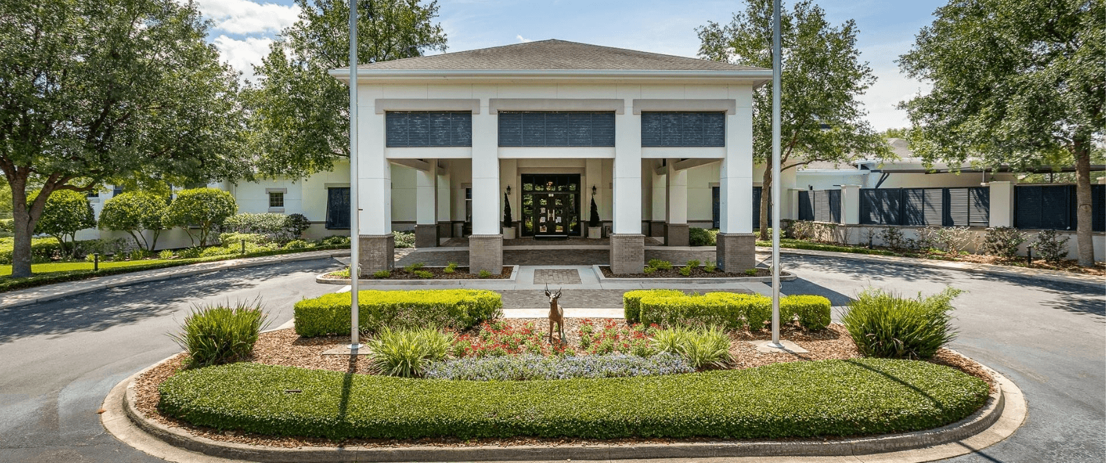 White building with columns, surrounded by greenery and circular driveway. Blue sky.