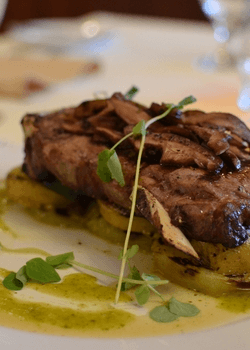Steak with mushrooms on potato slices, garnished with green sauce and sprigs, on a white plate.