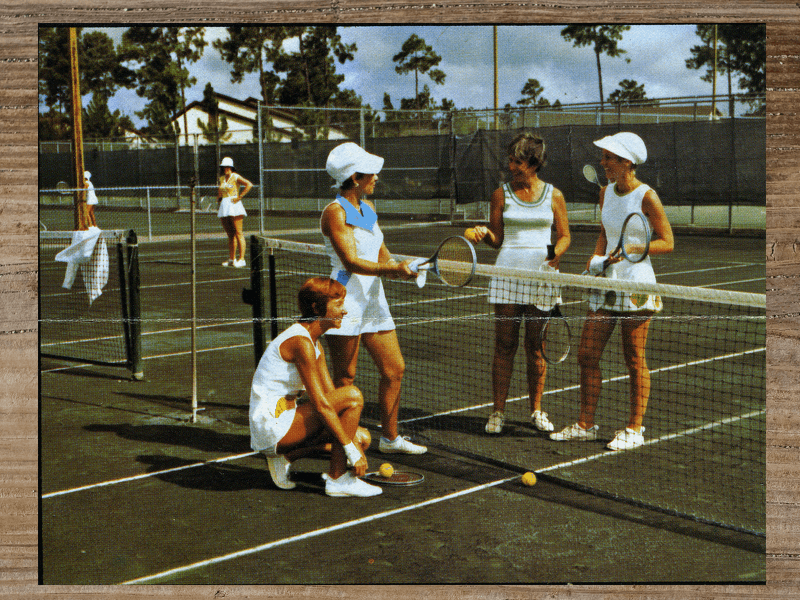 Women in tennis outfits on a green court, holding racquets and talking near the net.