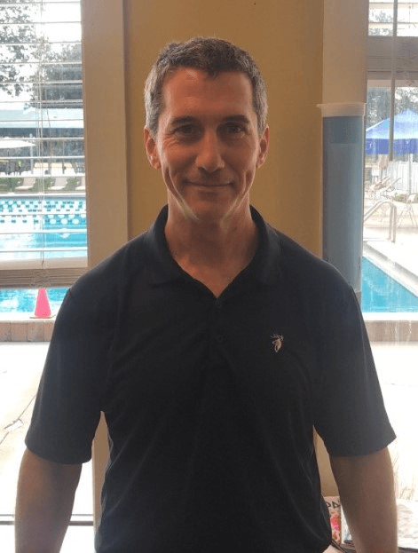 Tennis player in white shorts and visor swings racket on green court, shadow visible.