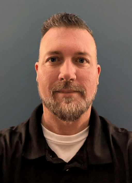 Tennis player in white shorts and visor swings racket on green court, shadow visible.