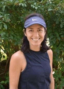 Tennis player in white shorts and visor swings racket on green court, shadow visible.