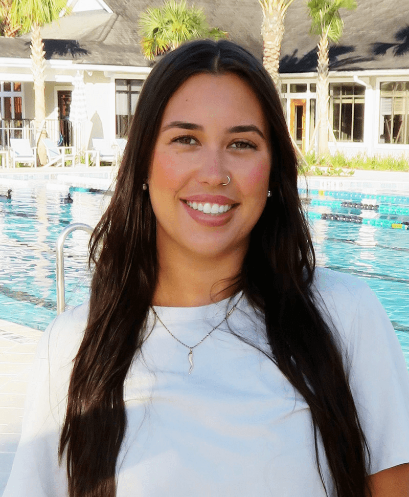 Tennis player in white shorts and visor swings racket on green court, shadow visible.
