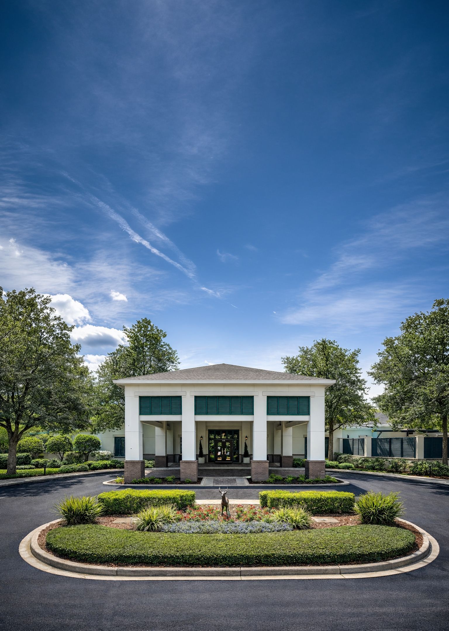 White building with columns, surrounded by greenery and circular driveway. Blue sky.