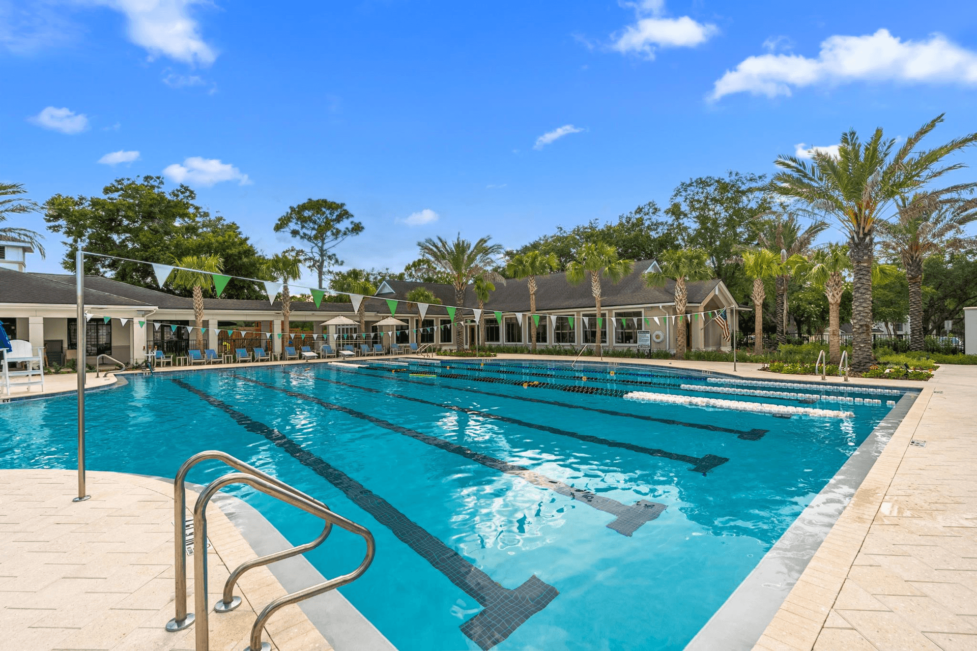 Outdoor swimming pool with lane markers, palm trees, and a clubhouse under a blue sky.
