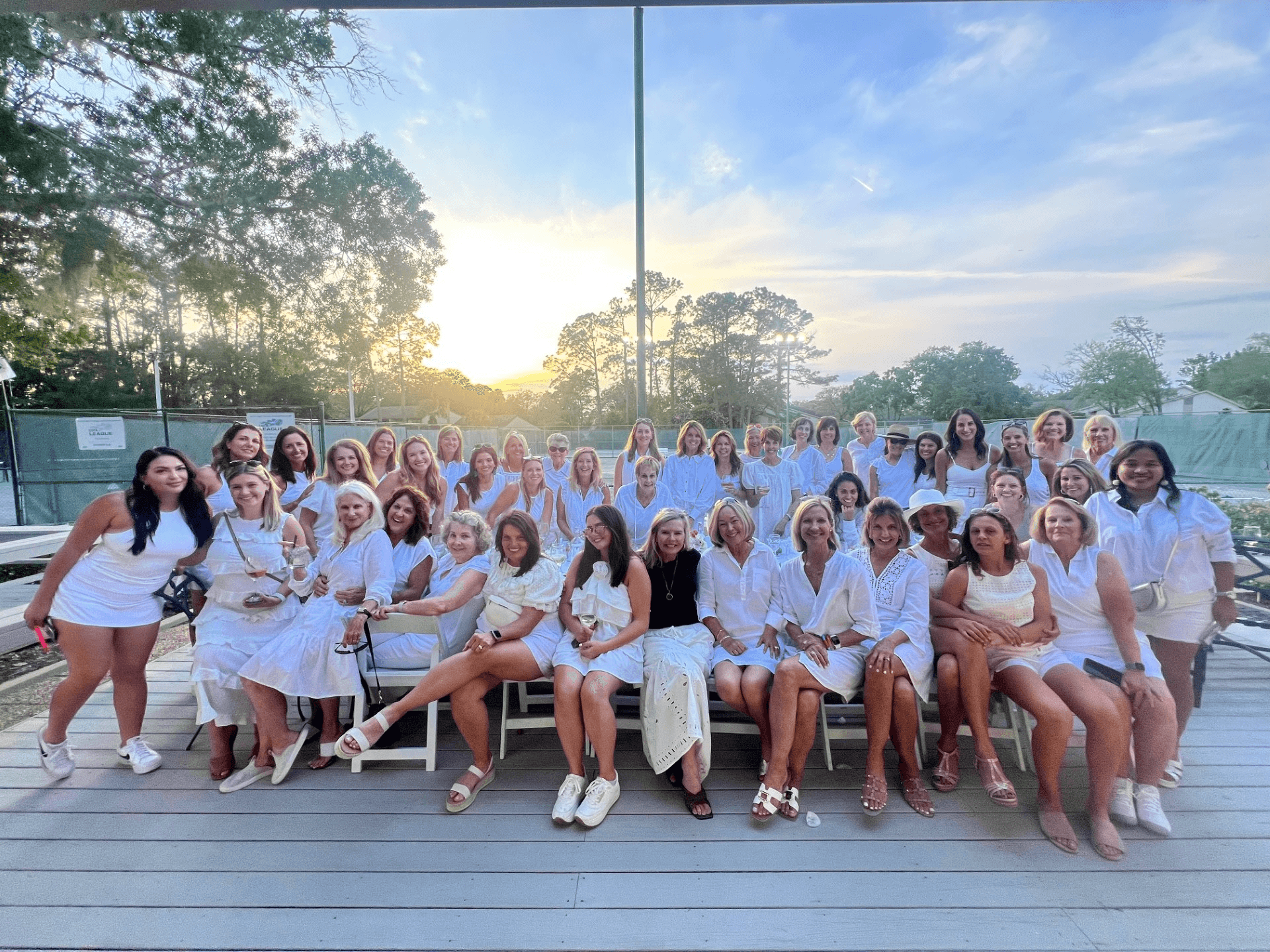 Group of people in white clothing, posing outdoors. Sunset background, wooden deck, trees visible.