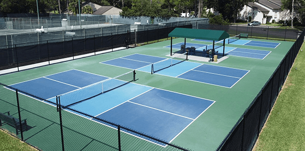 Outdoor pickleball courts, green and blue, surrounded by a fence, and a covered pavilion in the center.