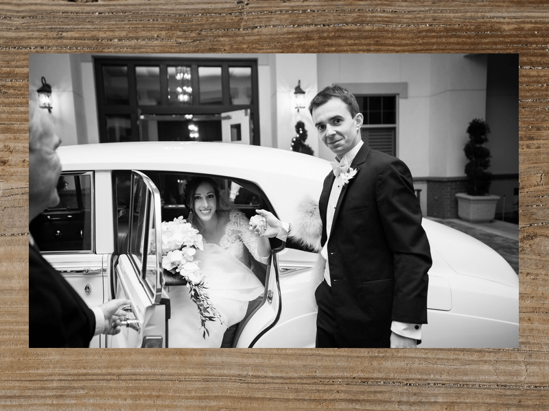 Bride exiting vintage car with groom assisting. Exterior shot with building in background, black and white.