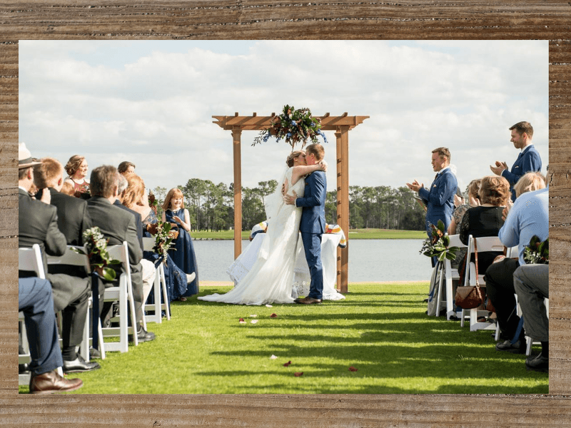 Wedding ceremony: Couple kissing under wooden arch, guests seated on either side, overlooking lake.