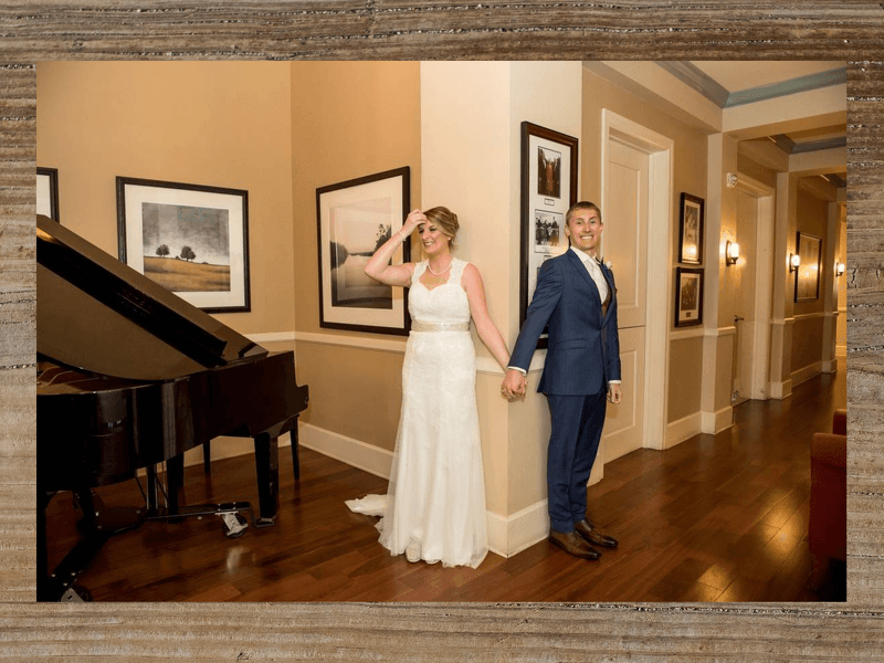 Bride and groom holding hands, smiling, in a hallway with a piano, paintings, and hardwood floor.