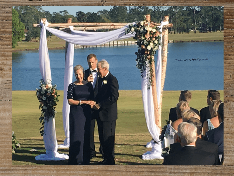 Wedding ceremony beside a lake: Couple under arch, officiant speaking, guests seated.