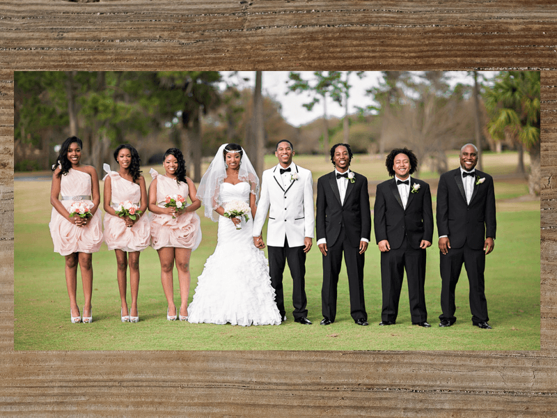Wedding party posing outdoors: bride in white gown, groom in white jacket, bridesmaids in pink dresses, groomsmen in black tuxedos.