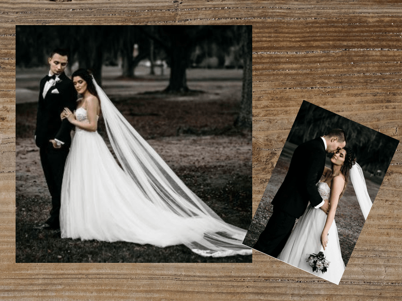 Wedding couple posing outdoors. Bride in white gown with long veil; groom in black suit. Photo on wood surface.