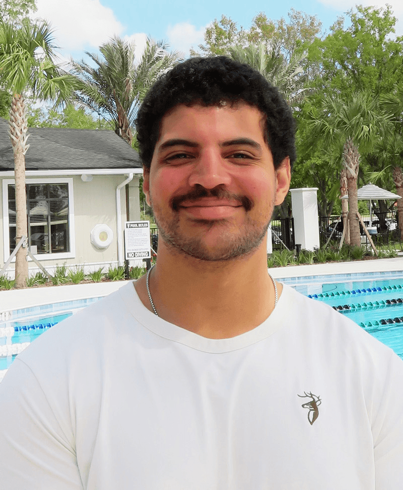 Tennis player in white shorts and visor swings racket on green court, shadow visible.