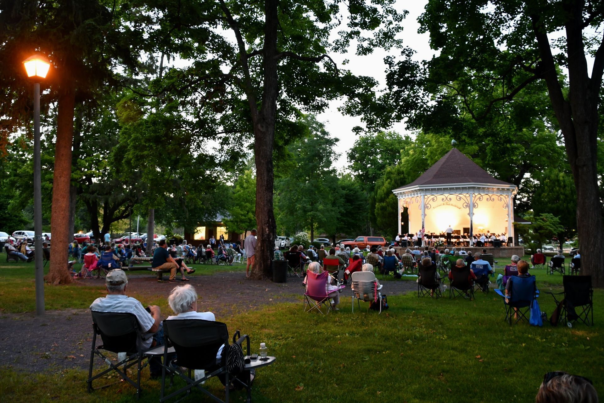 People seated in a park at dusk, watching a lit stage with trees and string lights nearby.