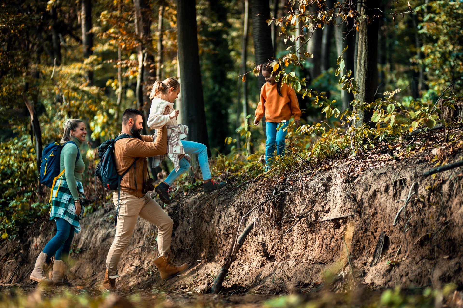 Four hikers walk through a wooded trail, one carrying a child, near a steep dirt embankment.