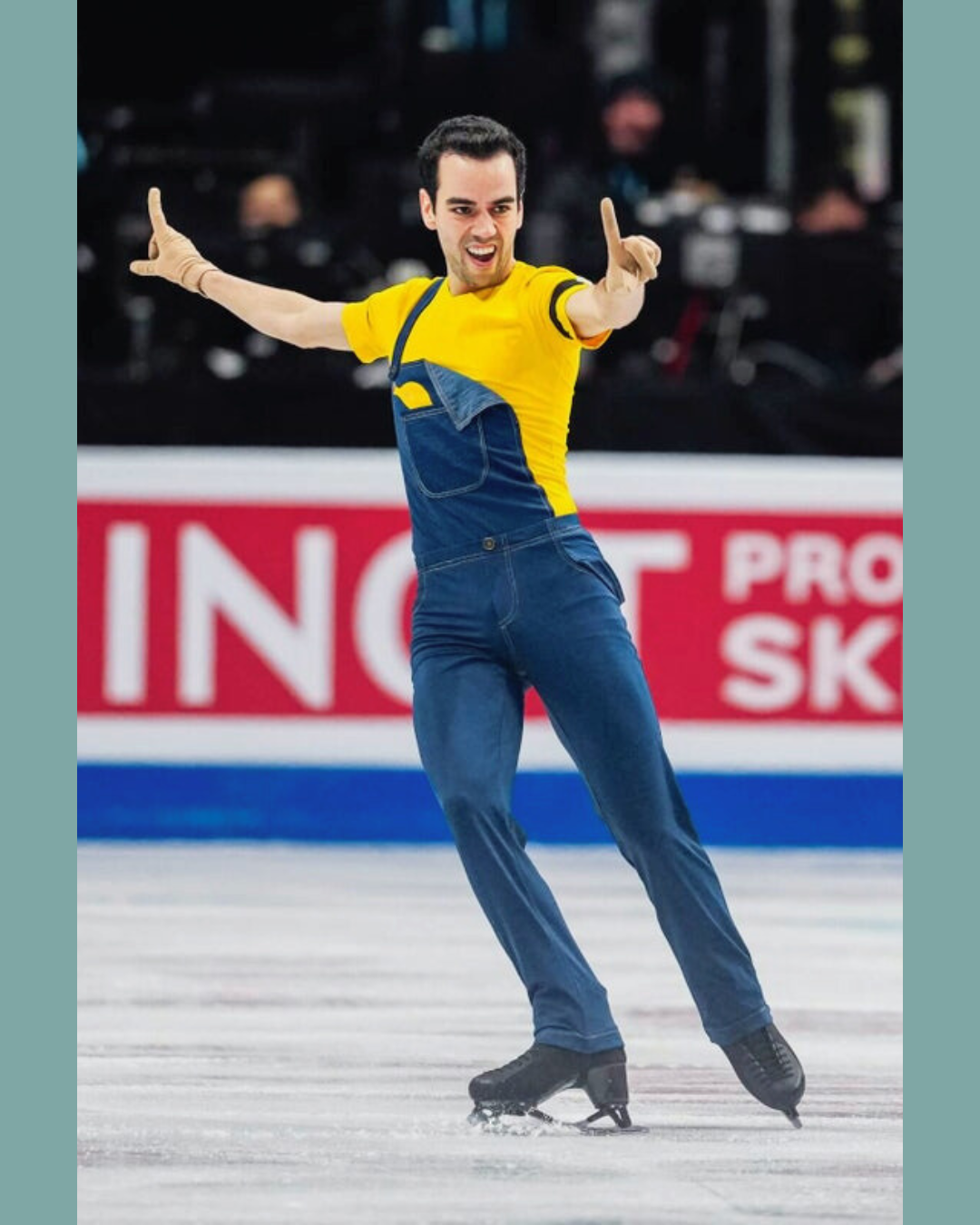 Figure skater in yellow and blue costume gliding on ice with arms outstretched, in front of rink boards