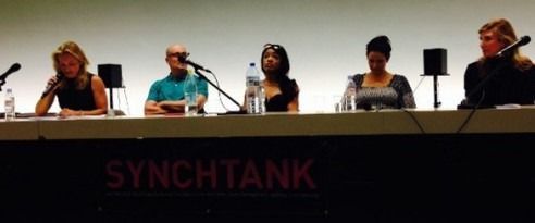 Five speakers seated at a Synchtank panel table on stage with microphones